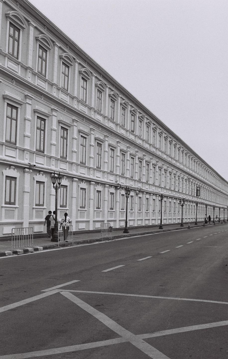A long, multi-story building with many windows lines a street with a few people walking on the sidewalk.