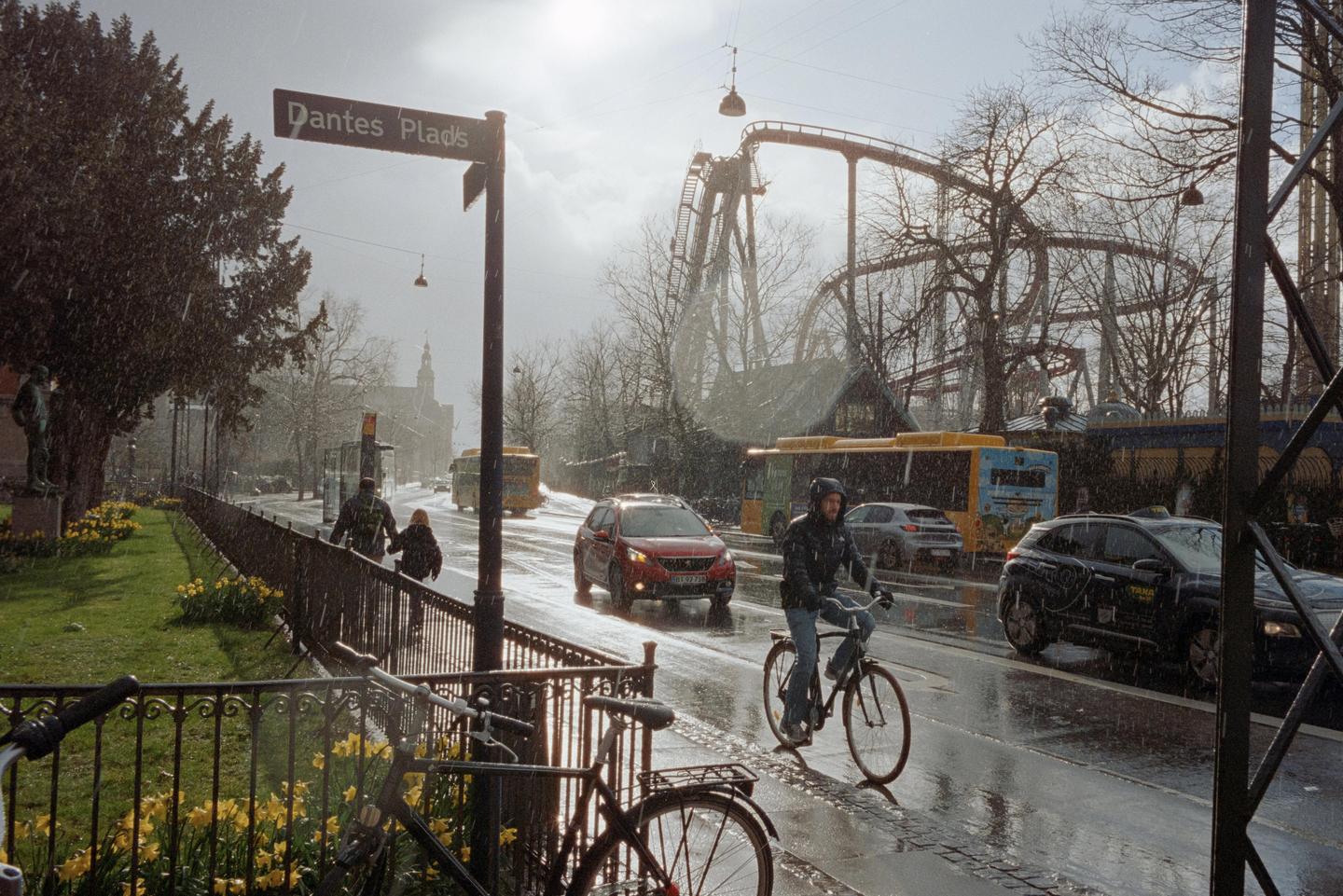 A street sign for Dantes Plads stands on a rainy street with cars, buses, and a cyclist, with a roller coaster in the background.