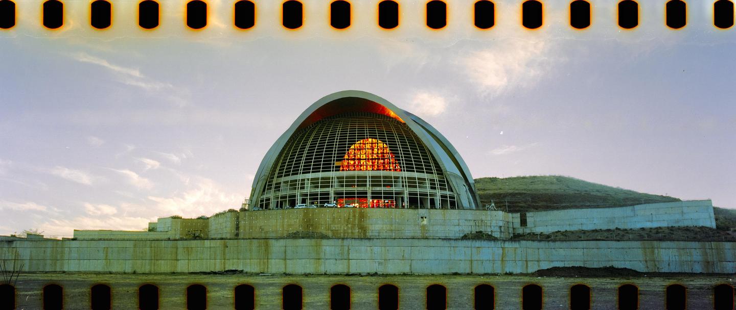 A large domed building with a grid-like facade and glowing orange windows sits on a concrete base next to a grassy hill under a cloudy sky.