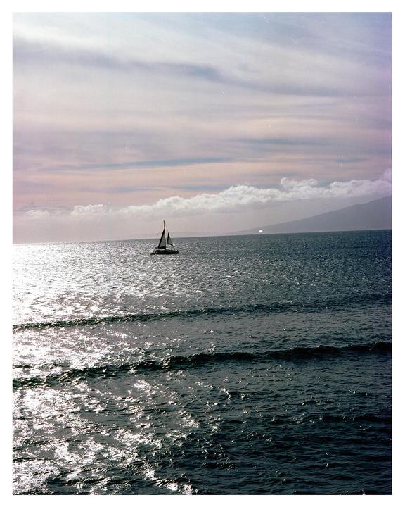 A sailboat is visible on the ocean with a mountain in the distance under a cloudy sky.