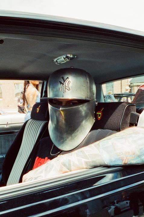 A man wearing a metallic helmet and a baseball cap is sitting in a car.