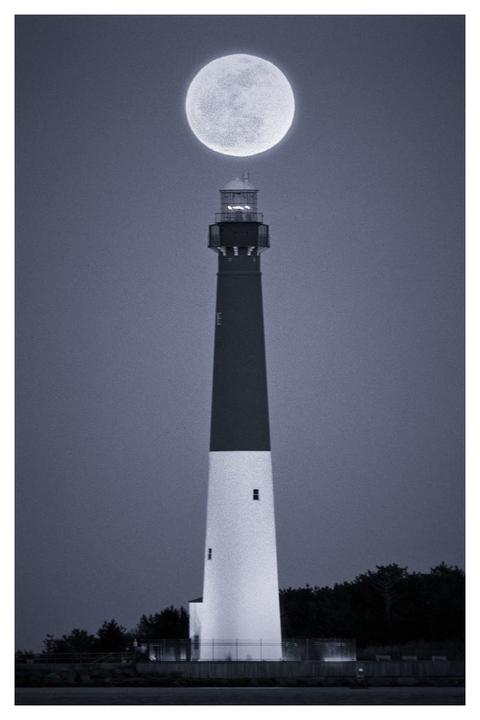 A full moon is positioned directly above a tall lighthouse at night.