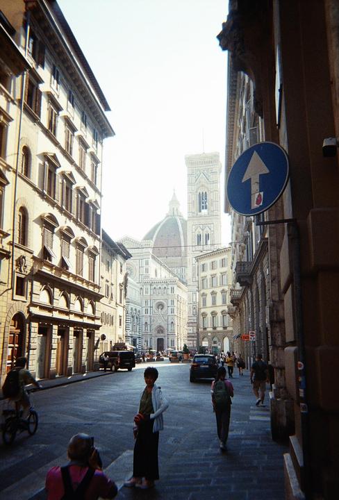 A street in Florence leads to the Duomo, with people walking and a blue arrow sign on the right.