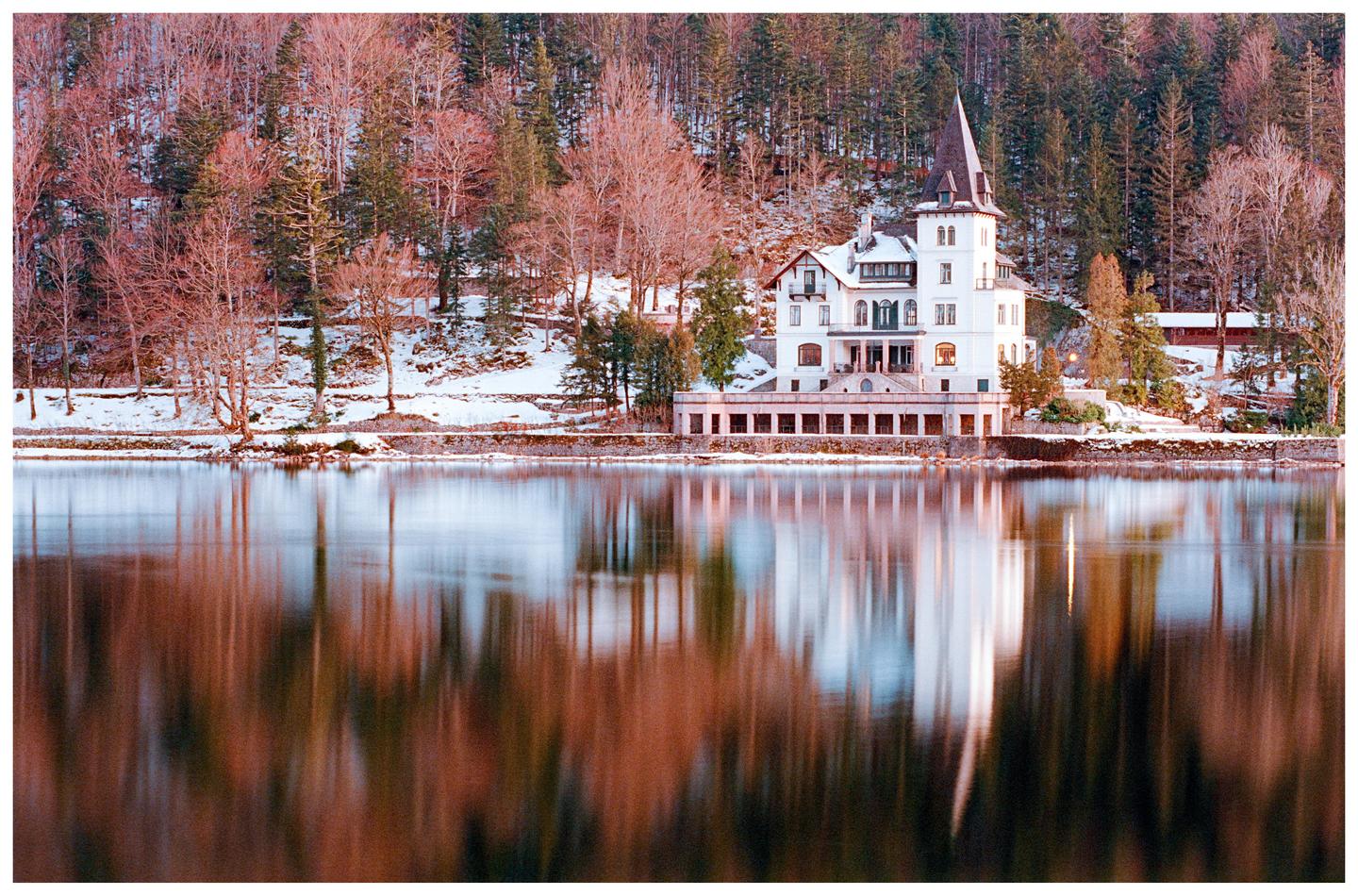 A white building with a pointed roof stands by a lake with a snow-covered forest in the background.