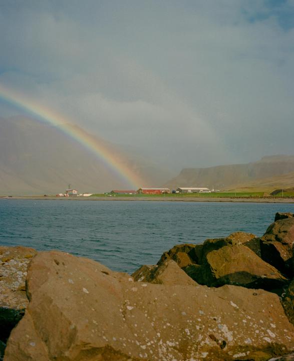 A rainbow stretches across a cloudy sky over a body of water with buildings on the distant shore and rocks in the foreground.