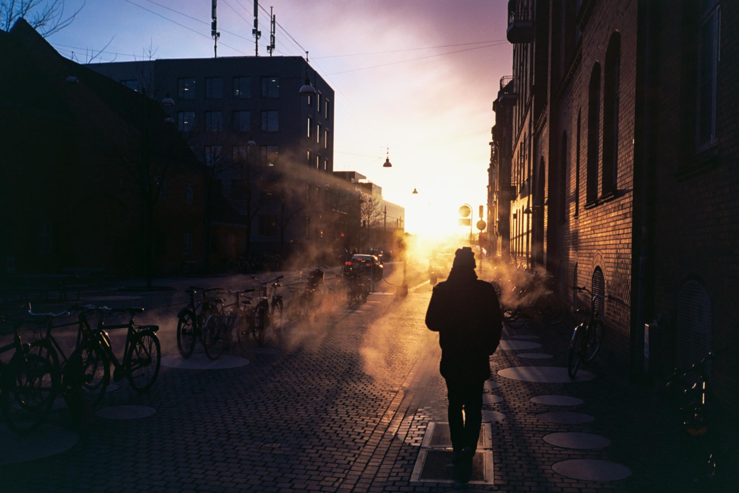 A person walks down a cobblestone street with parked bicycles as the sun sets, casting long shadows and illuminating steam rising from the ground.