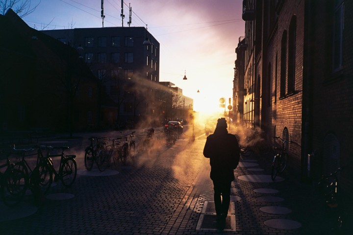 A person walks down a cobblestone street with parked bicycles as the sun sets, casting long shadows and illuminating steam rising from the ground.