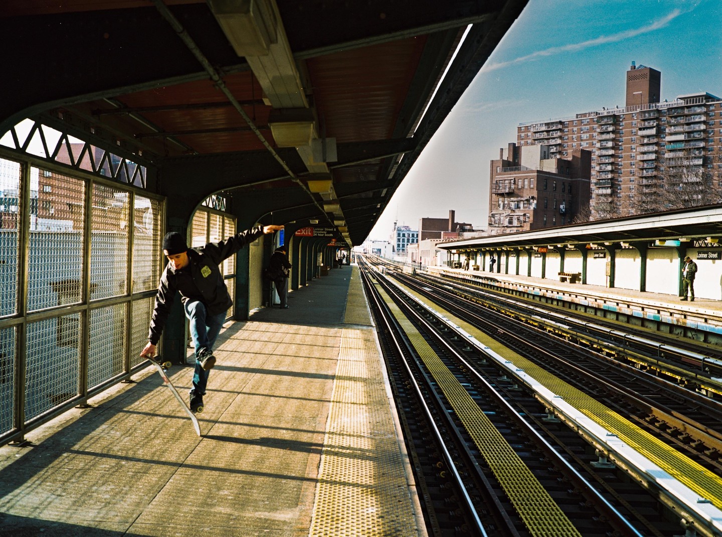 A man balances on a skateboard on a subway platform next to train tracks and buildings.