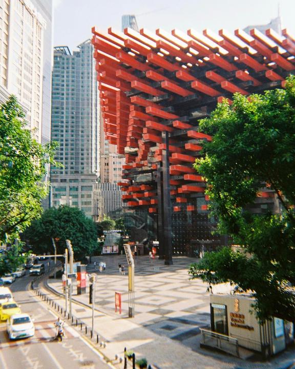 A modern building with red horizontal beams is visible next to a street with cars and trees.