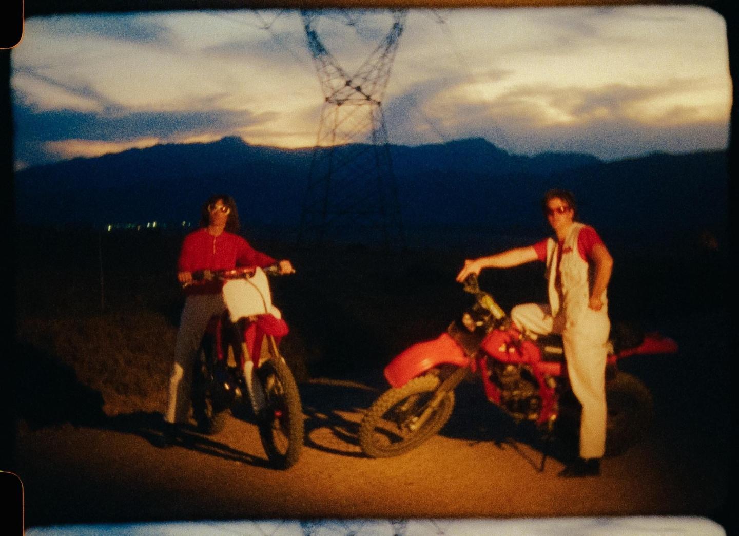 Two men pose with dirt bikes at dusk with mountains and a power line tower in the background.