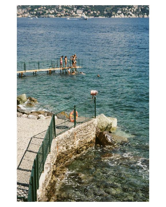 A group of people are swimming and standing on a pier in the sea with a town and hills in the background.