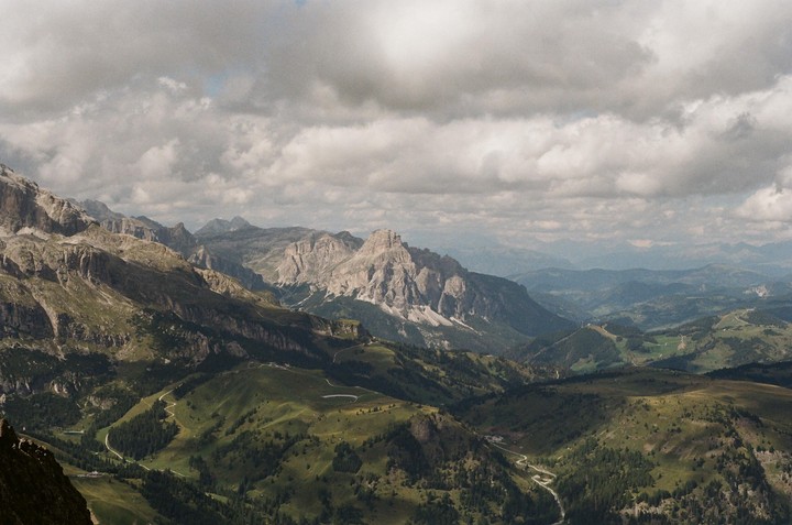 A winding road cuts through a lush green valley surrounded by towering mountains under a cloudy sky.