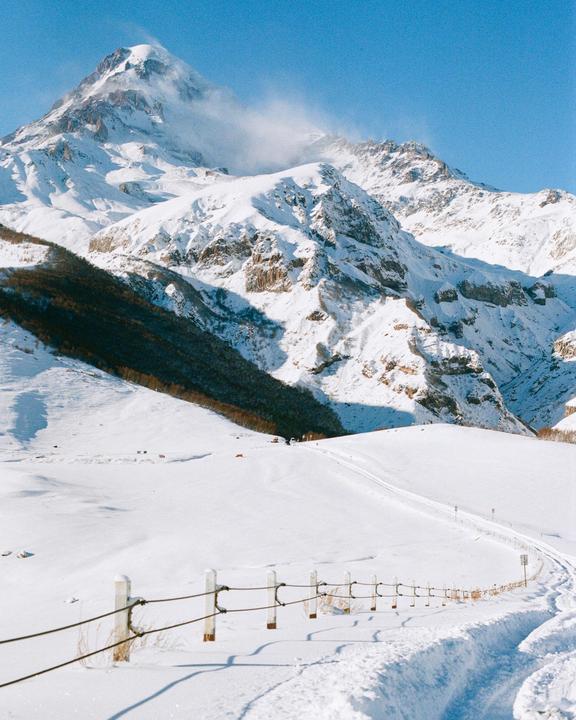 A snow-covered mountain range with a winding road and fence in the foreground is visible under a clear blue sky.