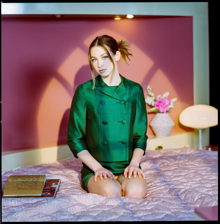 A woman in a green suit kneels on a pink quilted bedspread.