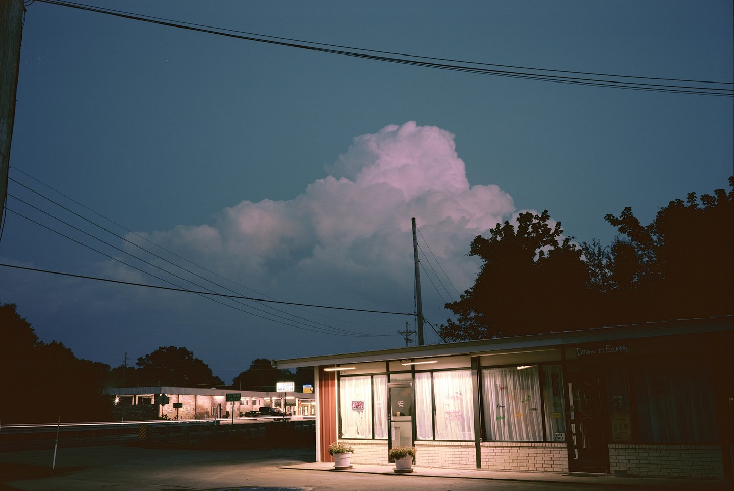A small building with illuminated windows and a sign that reads "Down to Earth" sits under a large, pink-tinged cloud at night.
