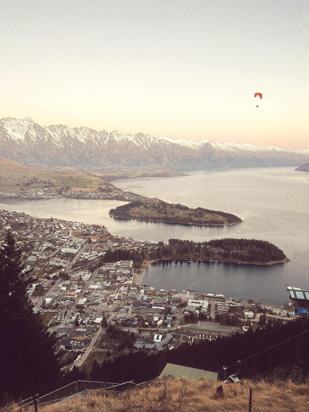 A paraglider flies over a town next to a lake with snow-capped mountains in the background.
