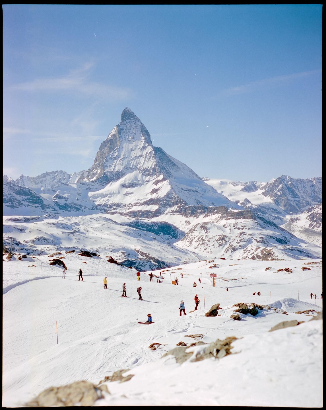 A wide shot shows a snow-covered mountain with skiers on a slope in the foreground.