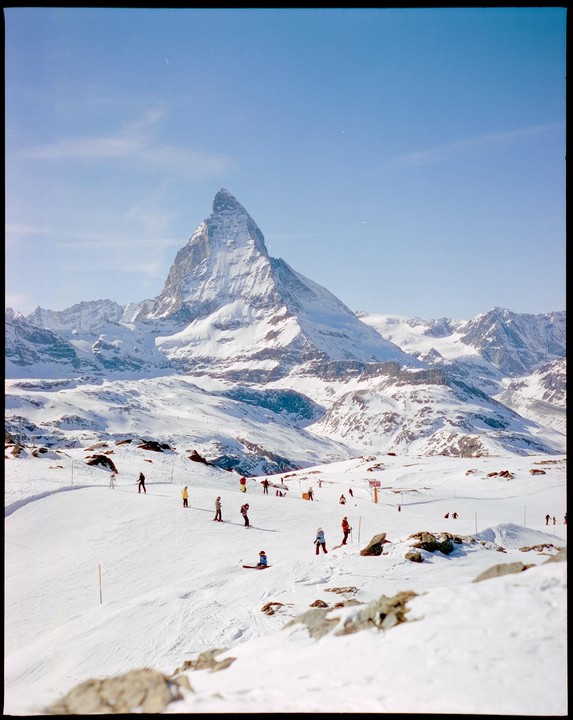A wide shot shows a snow-covered mountain with skiers on a slope in the foreground.
