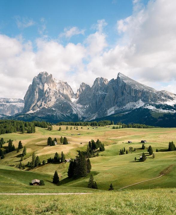 A vast green valley with scattered trees and small wooden huts stretches towards a range of snow-capped mountains under a partly cloudy sky.