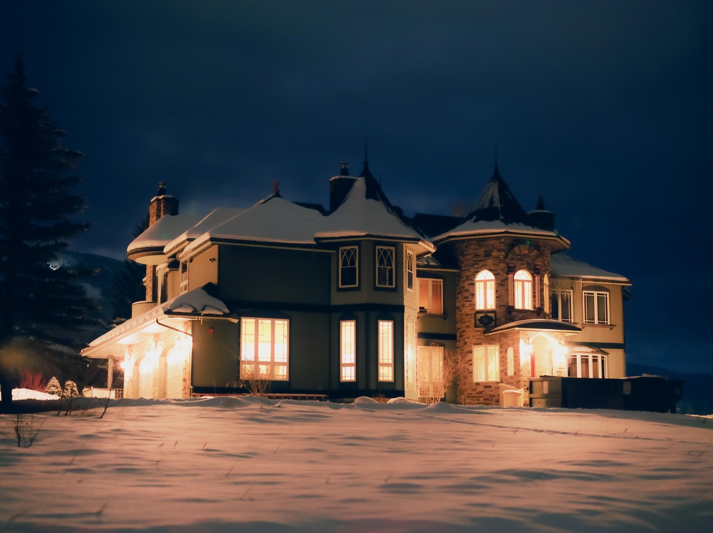 A large house with many windows and turrets is covered in snow at night.