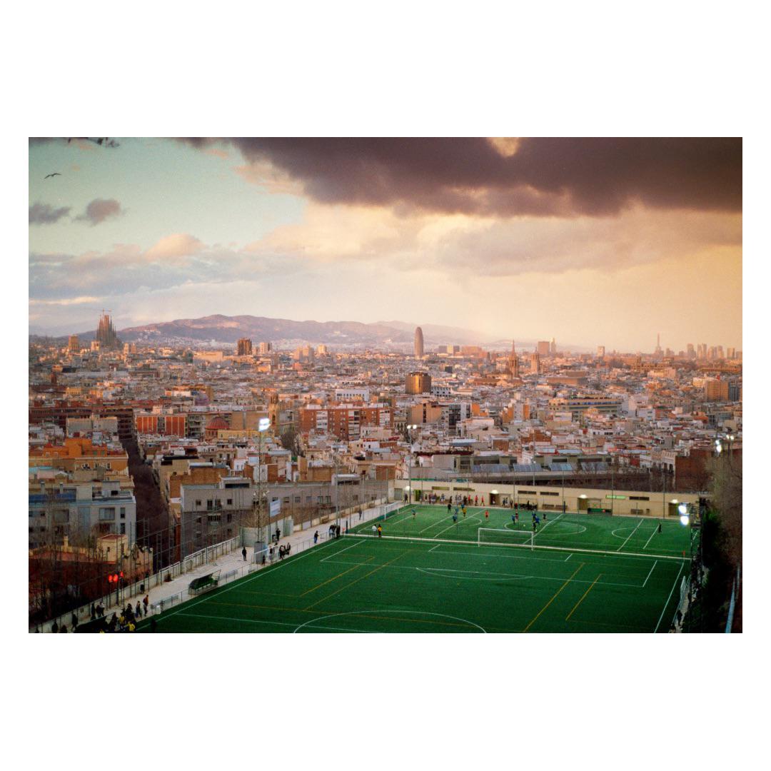 A city skyline with a soccer field in the foreground is visible under a cloudy sky at dusk.