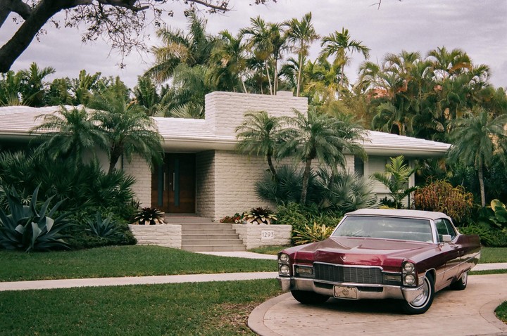 A vintage car is parked in the driveway of a mid-century modern house surrounded by palm trees.