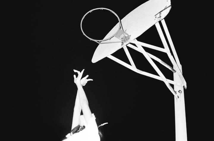 A person's arms are raised towards a basketball hoop and backboard against a dark background.