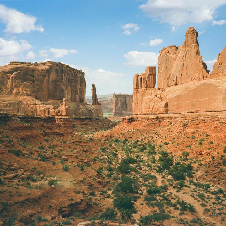 A wide shot shows a desert landscape with large red rock formations under a blue sky with white clouds.