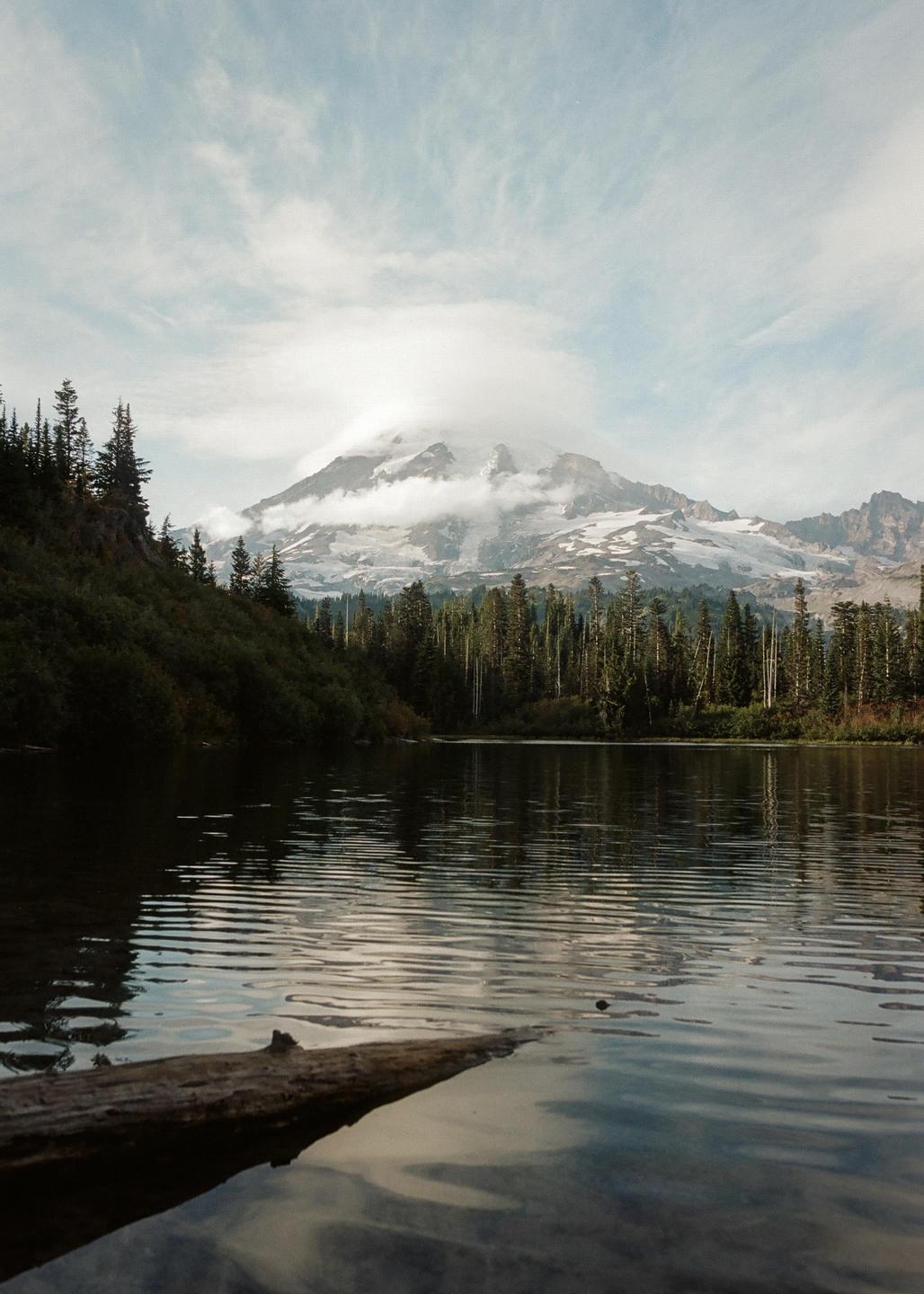 A snow-capped mountain with clouds around its peak rises above a forest and a lake with a log in the foreground.
