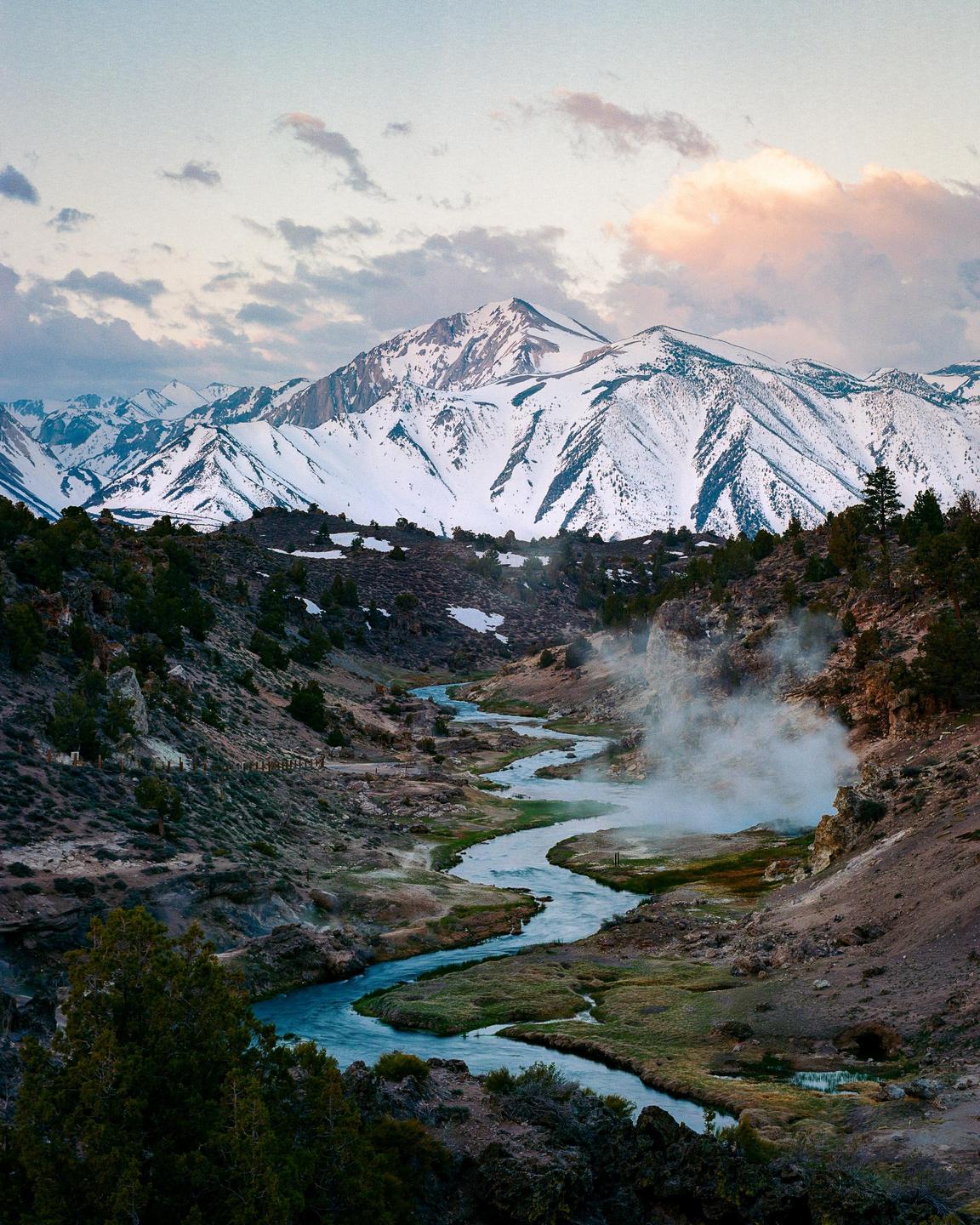 A winding river flows through a valley with steam rising from the bank, leading to snow-capped mountains under a cloudy sky.