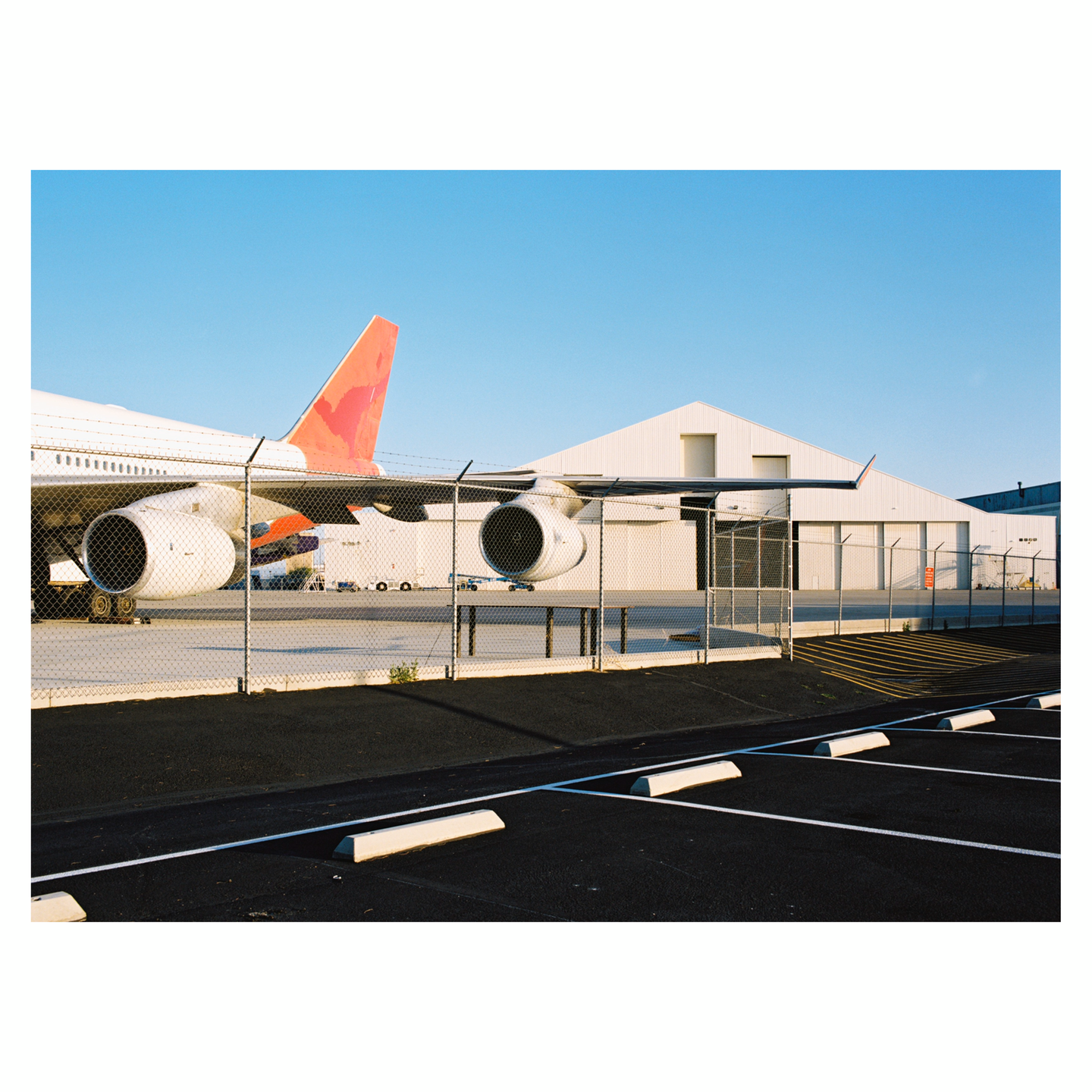 An airliner is parked behind a chain-link fence with a white hangar and an empty parking lot in the foreground.