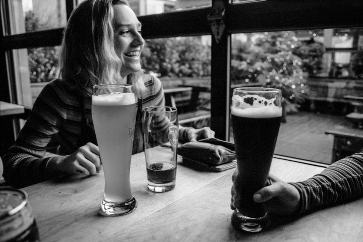 A woman smiles while sitting at a table with three glasses of beer, with a window looking out onto a decorated tree.