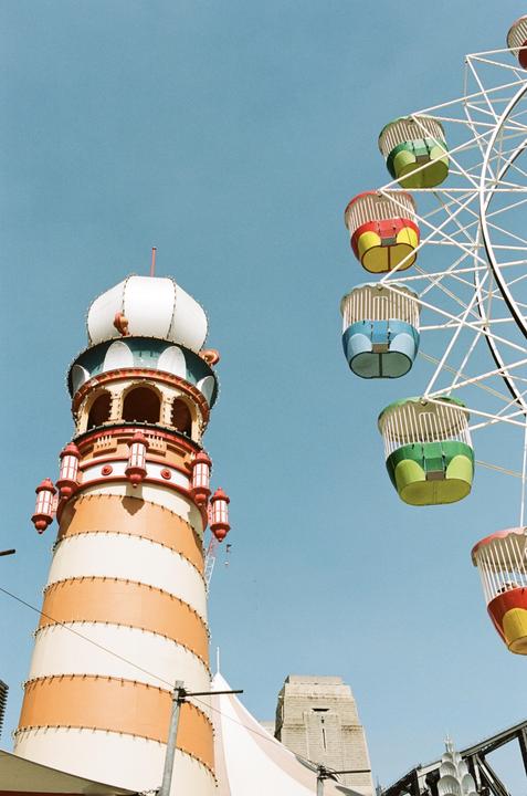 A tall, striped tower with a domed top stands next to a Ferris wheel with colorful gondolas against a clear blue sky.