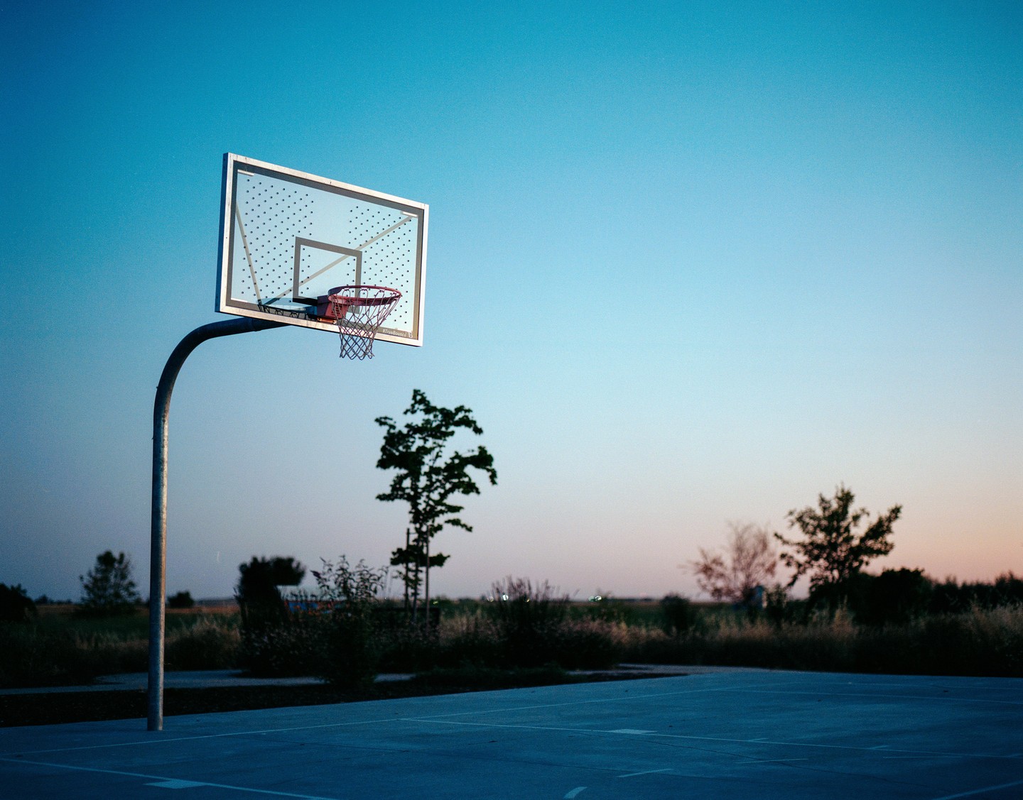 A basketball hoop stands on a court with trees and a field in the background under a clear sky at dusk.