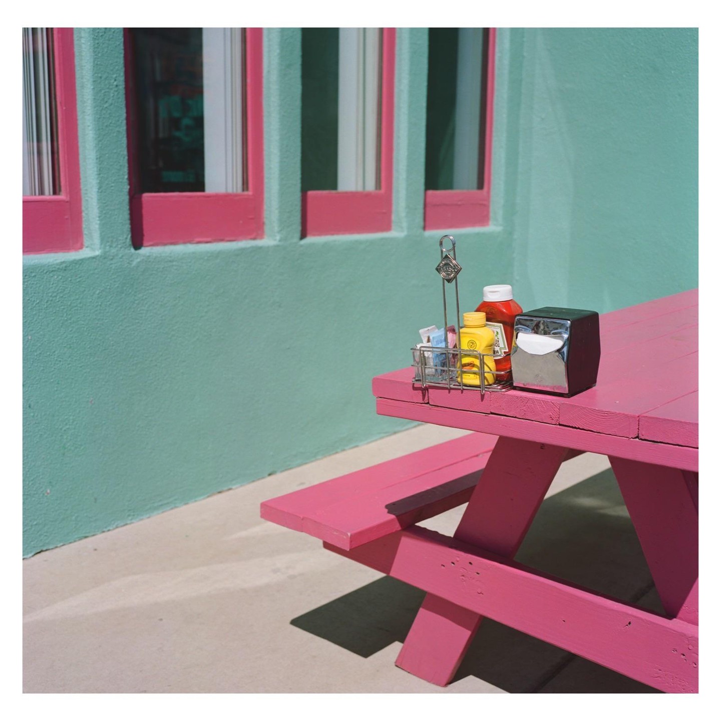 A pink picnic table with condiments sits outside a turquoise building with pink window frames.