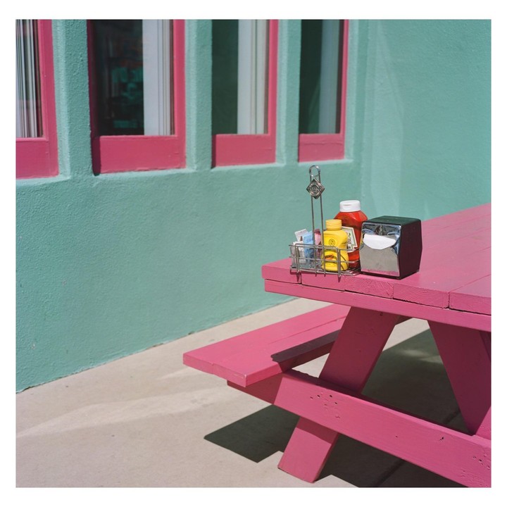 A pink picnic table with condiments sits outside a turquoise building with pink window frames.