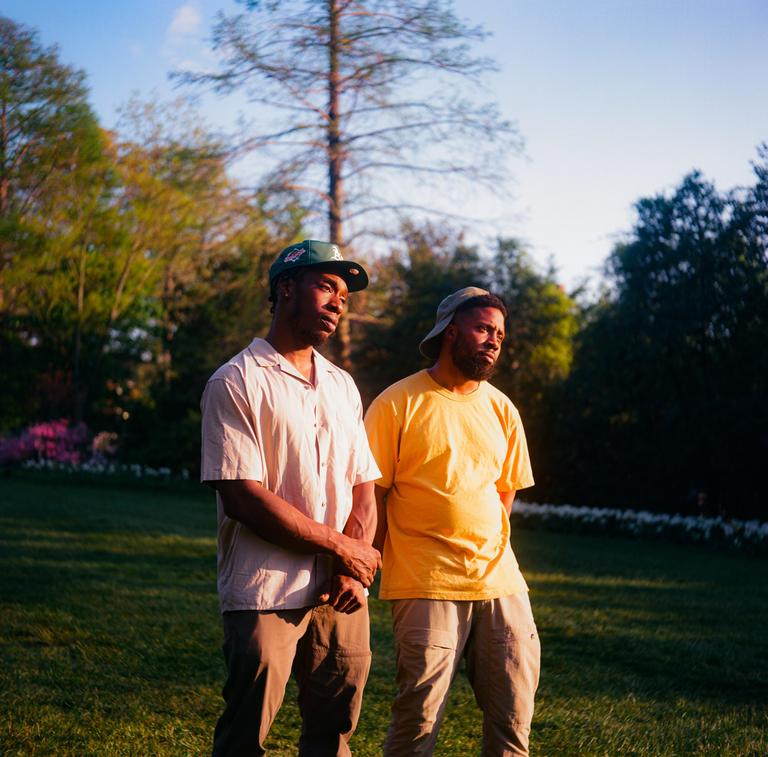 Two men stand in a grassy field with trees in the background under a bright sky.
