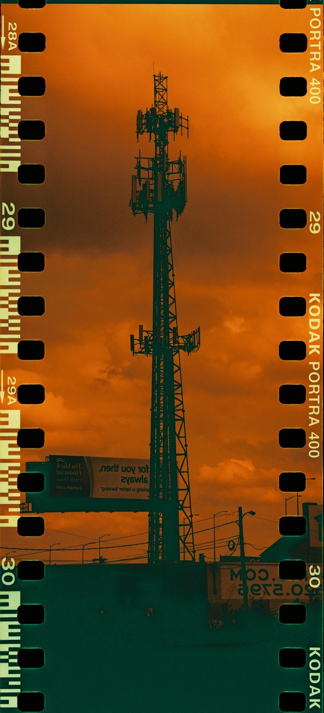 A tall communication tower stands silhouetted against an orange sky with clouds, with a billboard and power lines visible below.