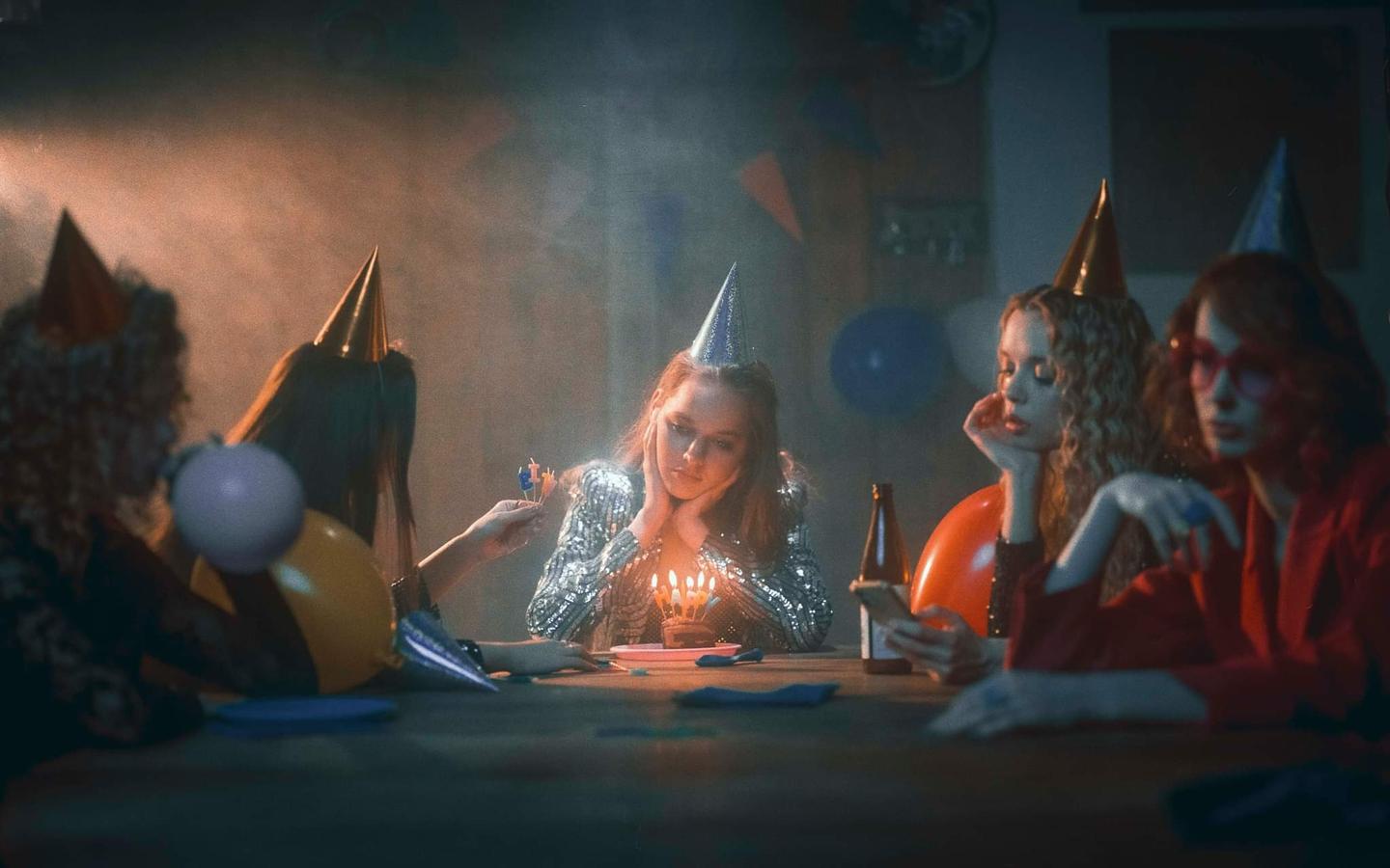 A group of women wearing party hats sit around a table with a birthday cake and lit candles.