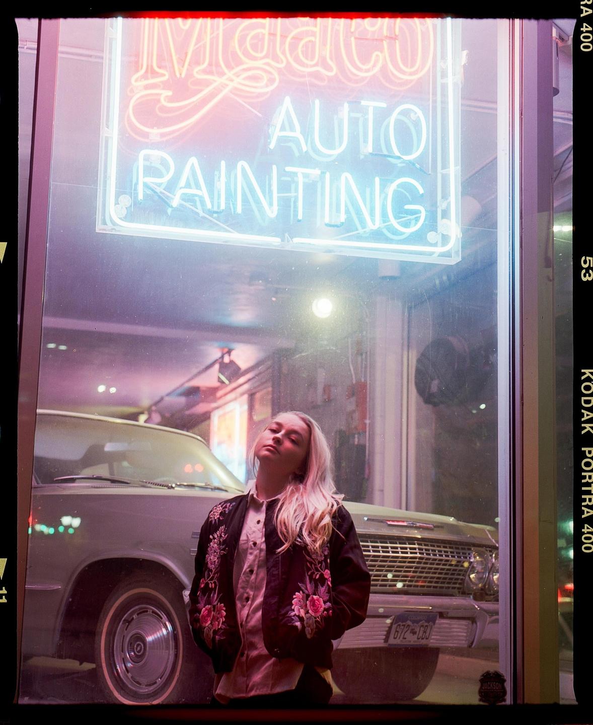 A woman with blonde hair stands in front of a car and a neon sign that reads "Auto Painting."