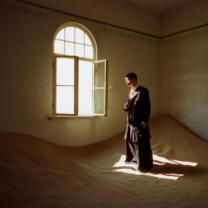 A man in a suit stands in a room filled with sand dunes, looking out an open window.
