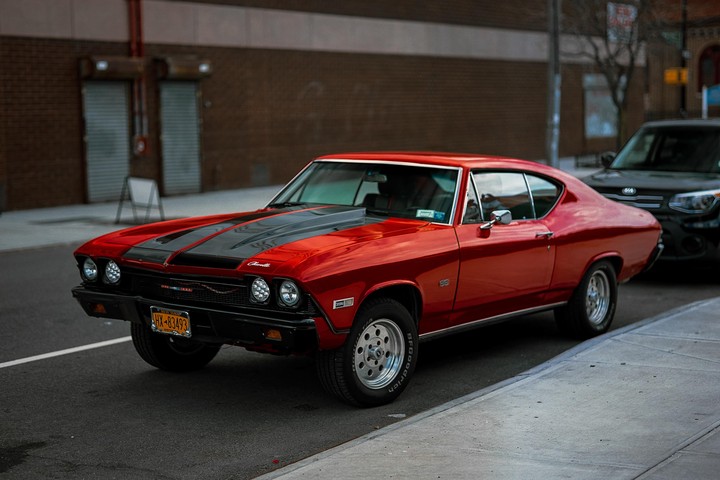 A red Chevelle with black racing stripes is parked on a city street next to a sidewalk.