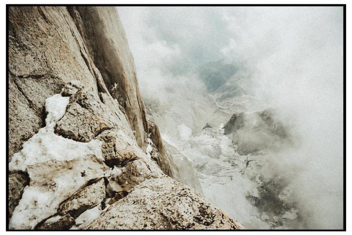 A steep, rocky mountain face with patches of snow descends into a valley filled with clouds.