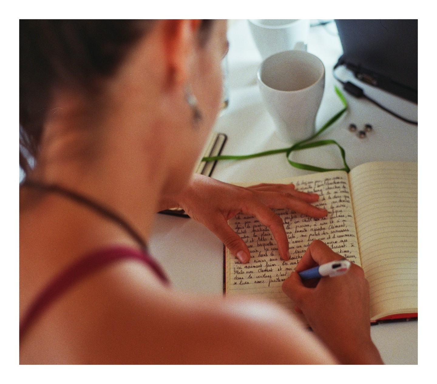 A woman writes in a notebook with a pen, with a white mug and a laptop visible in the background.