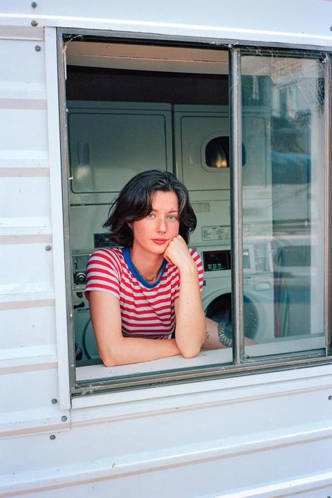 A woman in a striped shirt leans out of a window with washing machines visible in the background.