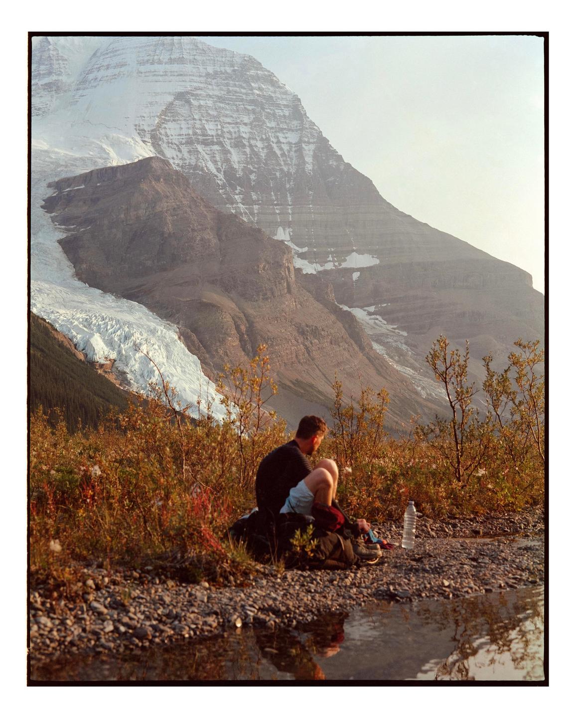 A man sits by a stream with a large snow-capped mountain in the background.