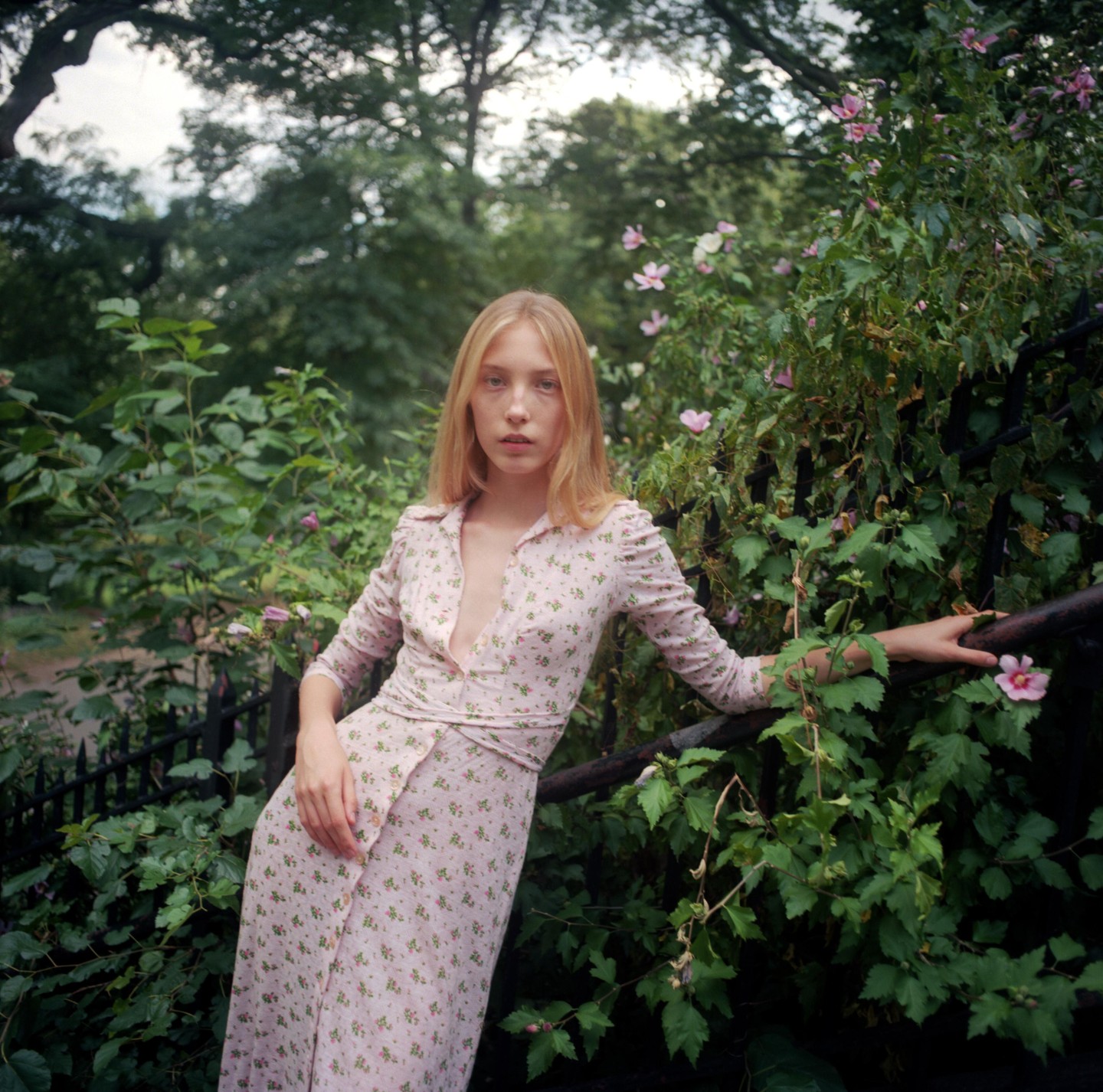 A woman in a floral dress leans against a fence covered in foliage and pink flowers.