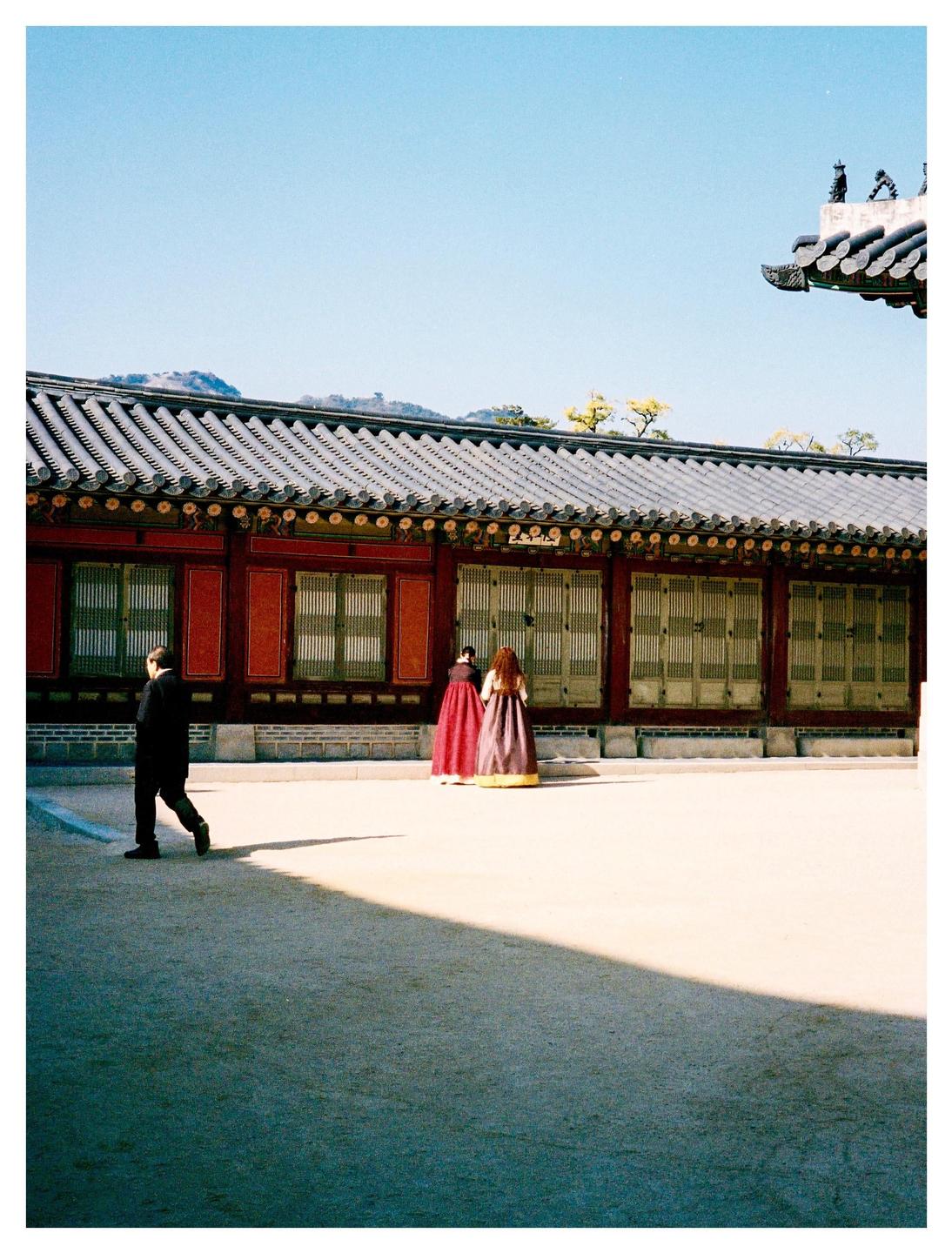 A man walks across a sunny courtyard while two women in traditional dress stand near a red building with a tiled roof.