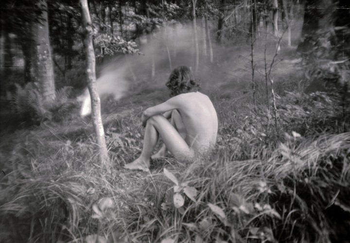 A nude person sits in tall grass in a forest with mist or smoke in the background.
