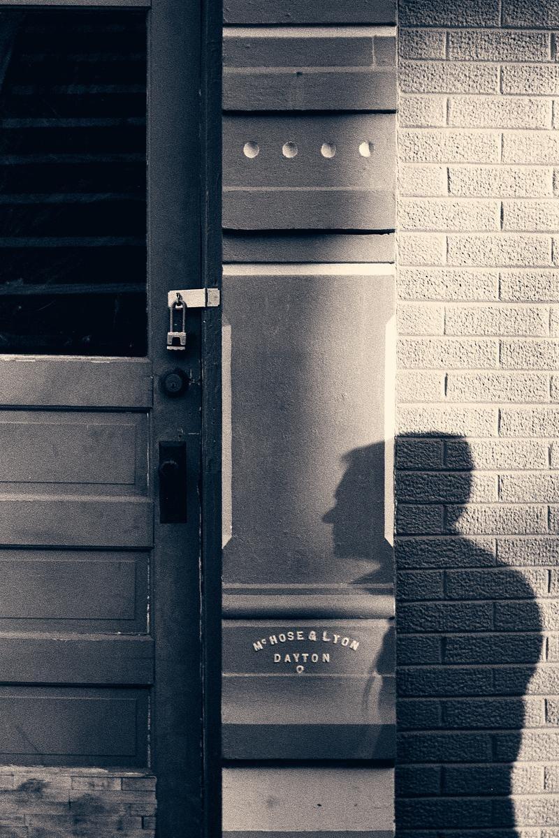 A shadow of a man is cast on a brick wall next to a door with a sign that reads "MC HOSE & LYON DAYTON".